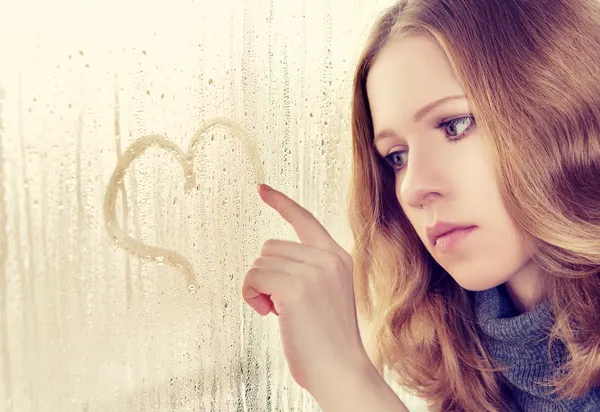 Sad girl draws a heart on the window in the rain — стоковое фото