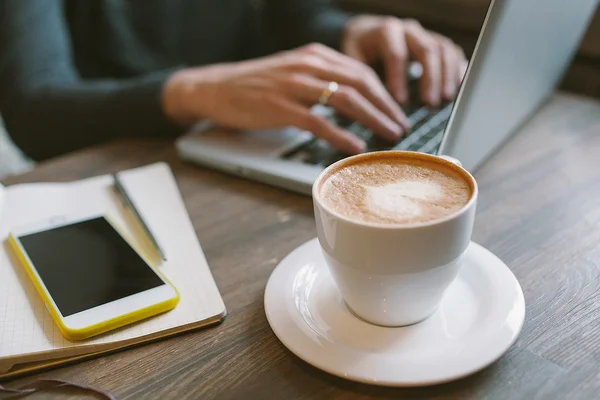 Руки человека на ноутбуке с кофе и смартфоне с блокнотом — стоковое фото